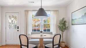 Comedor con mesa redonda, dos sillas, lámpara colgante, planta en maceta y ventana con cortinas.