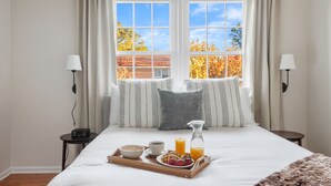 Una cama bien hecha con una bandeja de desayuno, una mesita de noche con una lámpara y una ventana con cortinas.