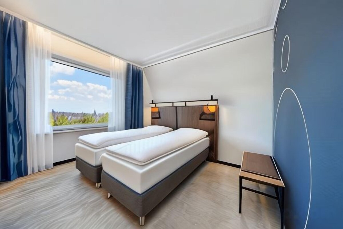 Una habitación de hotel con dos camas, un ventanal con cortinas y vistas al exterior, además de una pared con diseño geométrico.