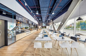 Un café moderno con una barra alargada, sillas blancas y ventanas amplias.