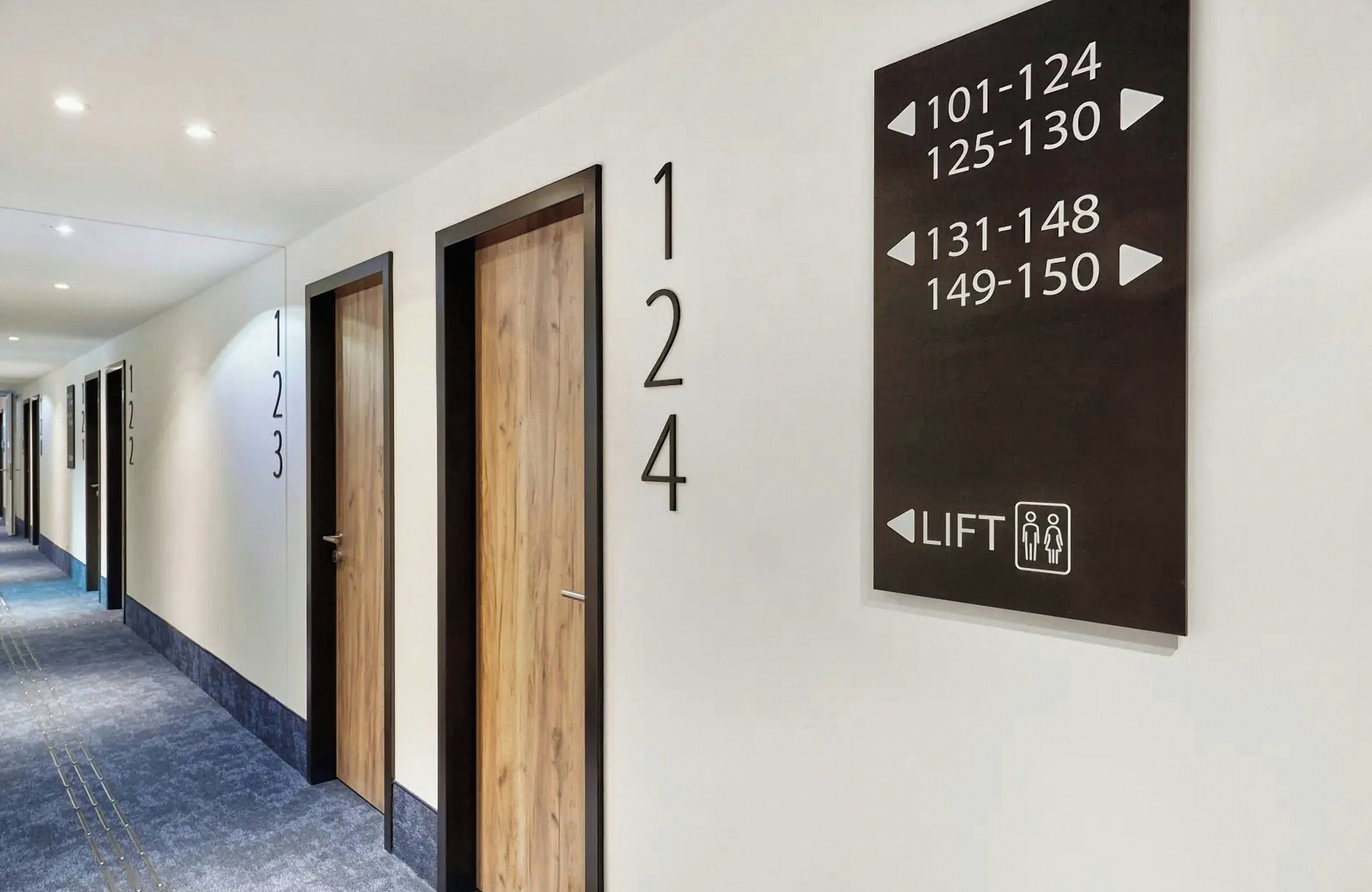 Un corridoio d'albergo con porte numerate, un cartello che indica la direzione degli ascensori e un pavimento rivestito di moquette.