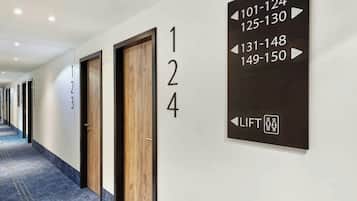 Un corridoio d'albergo con porte numerate, un cartello che indica la direzione degli ascensori e un pavimento rivestito di moquette.