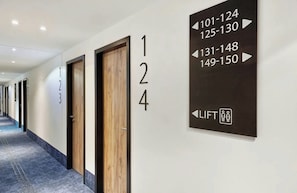 Pasillo de hotel con puertas numeradas, un indicador de dirección de ascensor y suelo alfombrado.