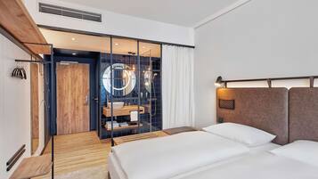 Una habitación de hotel moderna con piso de madera, una cama grande con dos almohadas, una mesita de noche y un espejo circular integrado.