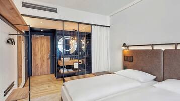 A modern hotel room with a wooden floor, a bed with white linens, a large mirror, and a bathroom visible through a glass partition.