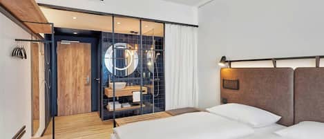A modern hotel room with a wooden floor, a bed with white linens, a large mirror, and a bathroom visible through a glass partition.