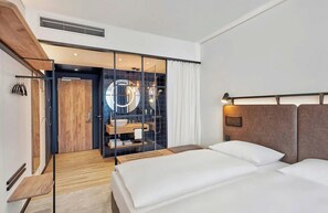 A modern hotel room with a wooden floor, a bed with white linens, a large mirror, and a bathroom visible through a glass partition. - Leonardo Smart Hotel Düsseldorf (Düsseldorf)