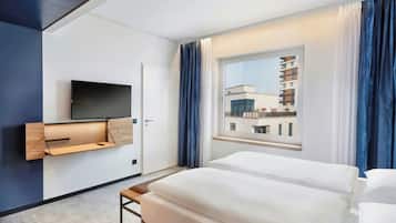 A hotel room with a bed, a TV mounted on the wall, a window with curtains, and a view of the building outside.