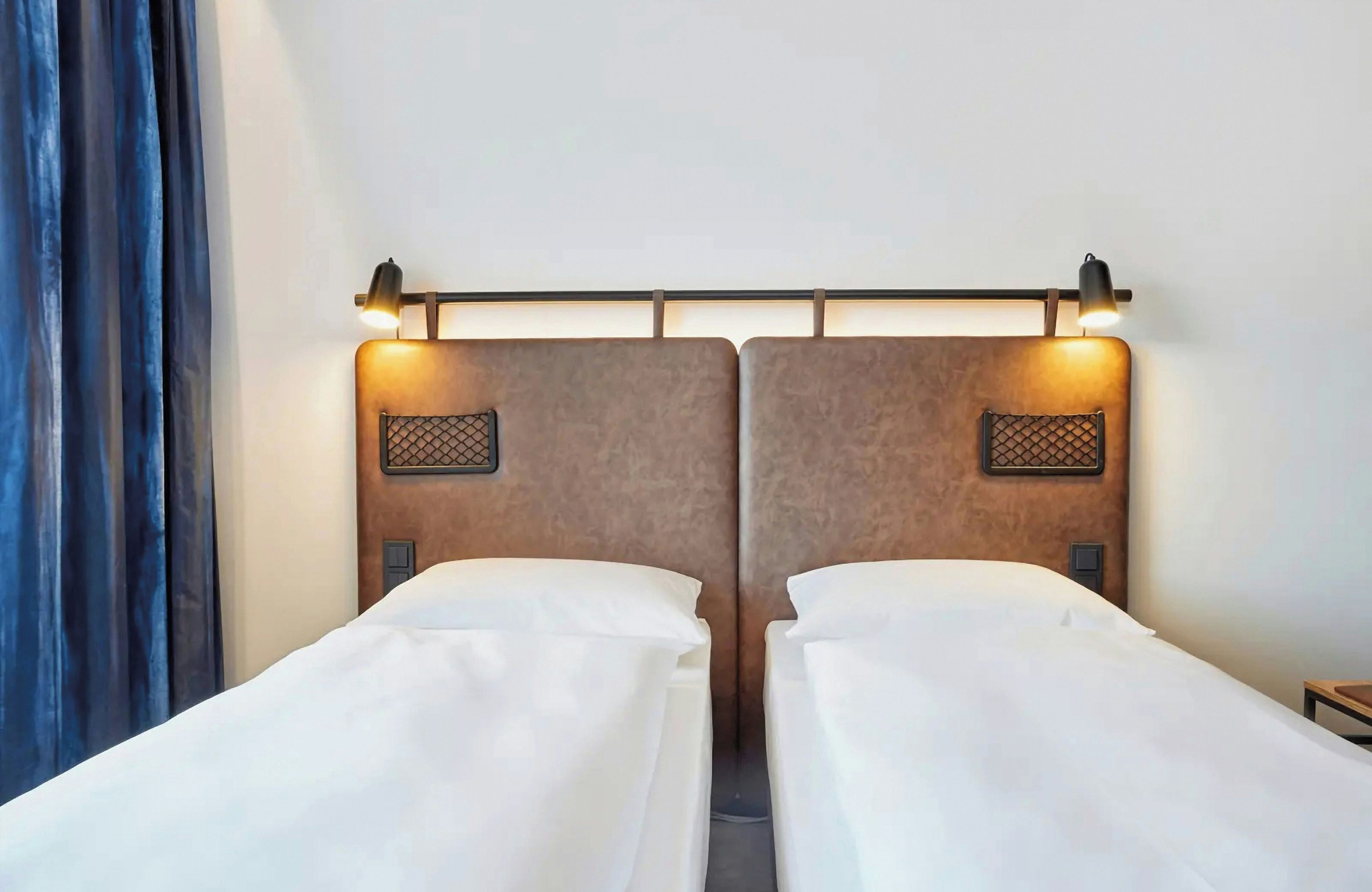 A hotel room with two single beds, a headboard with a light fixture, and a window with curtains.