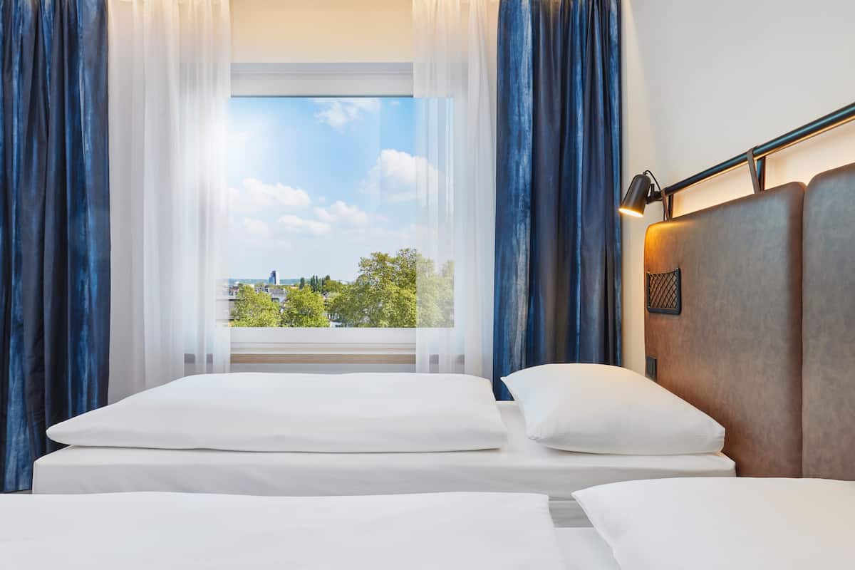 Ett hotellrum med ett stort fönster, en säng med vita sängkläder och en sänggavel med en lampa.