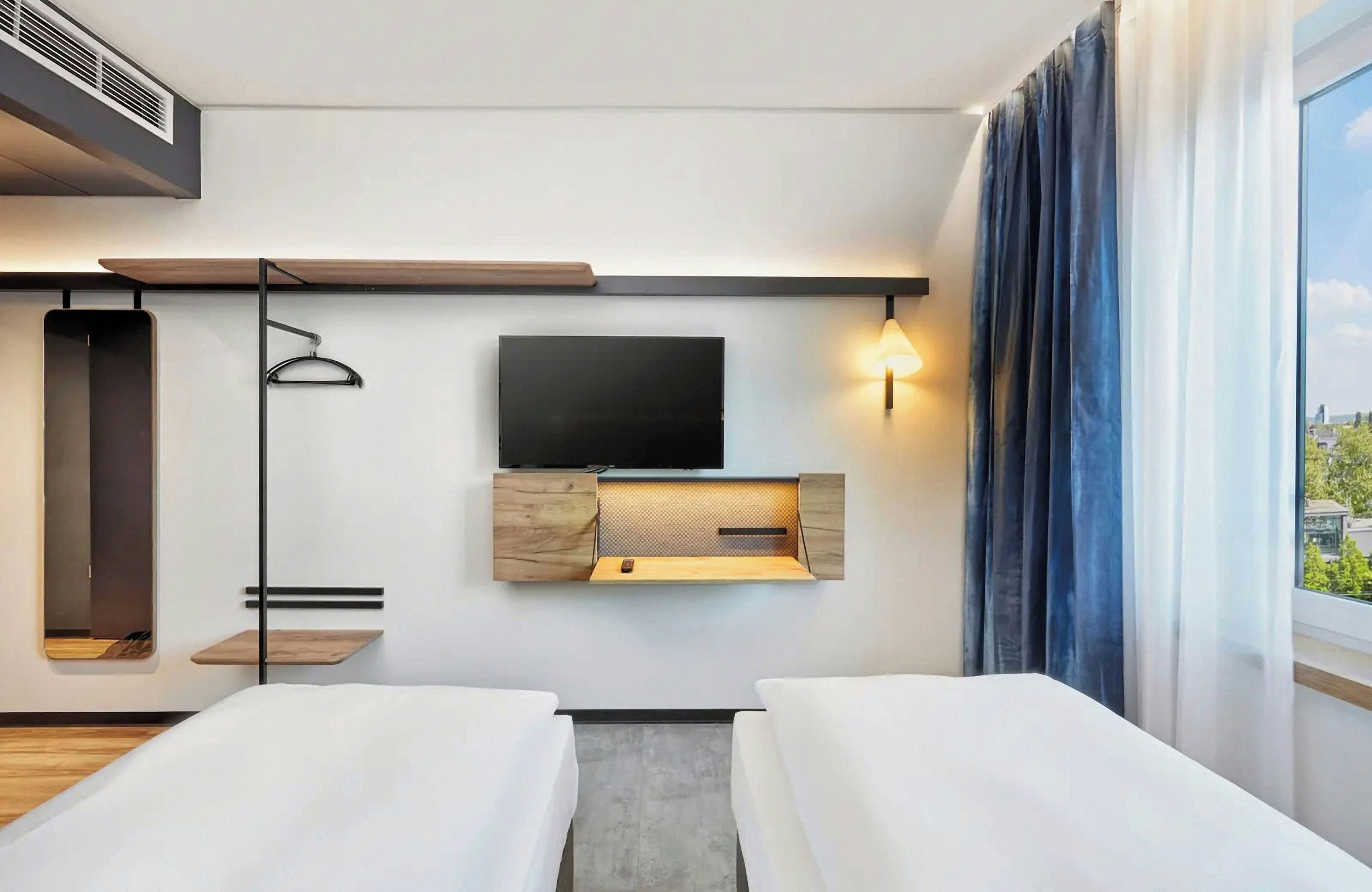 A modern hotel room with a flat-screen TV mounted on the wall, a bedside table with a lamp, and a large window with curtains.