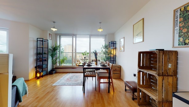 Un coin repas avec une table et des chaises en bois, une vue dégagée grâce à de grandes fenêtres et une bibliothèque ornée d’un tableau encadré au mur.