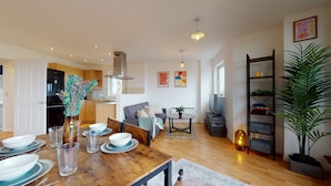 Une cuisine moderne avec des meubles en bois, un coin repas avec une table en bois dressée pour quatre personnes, et un salon avec un canapé et une table basse.