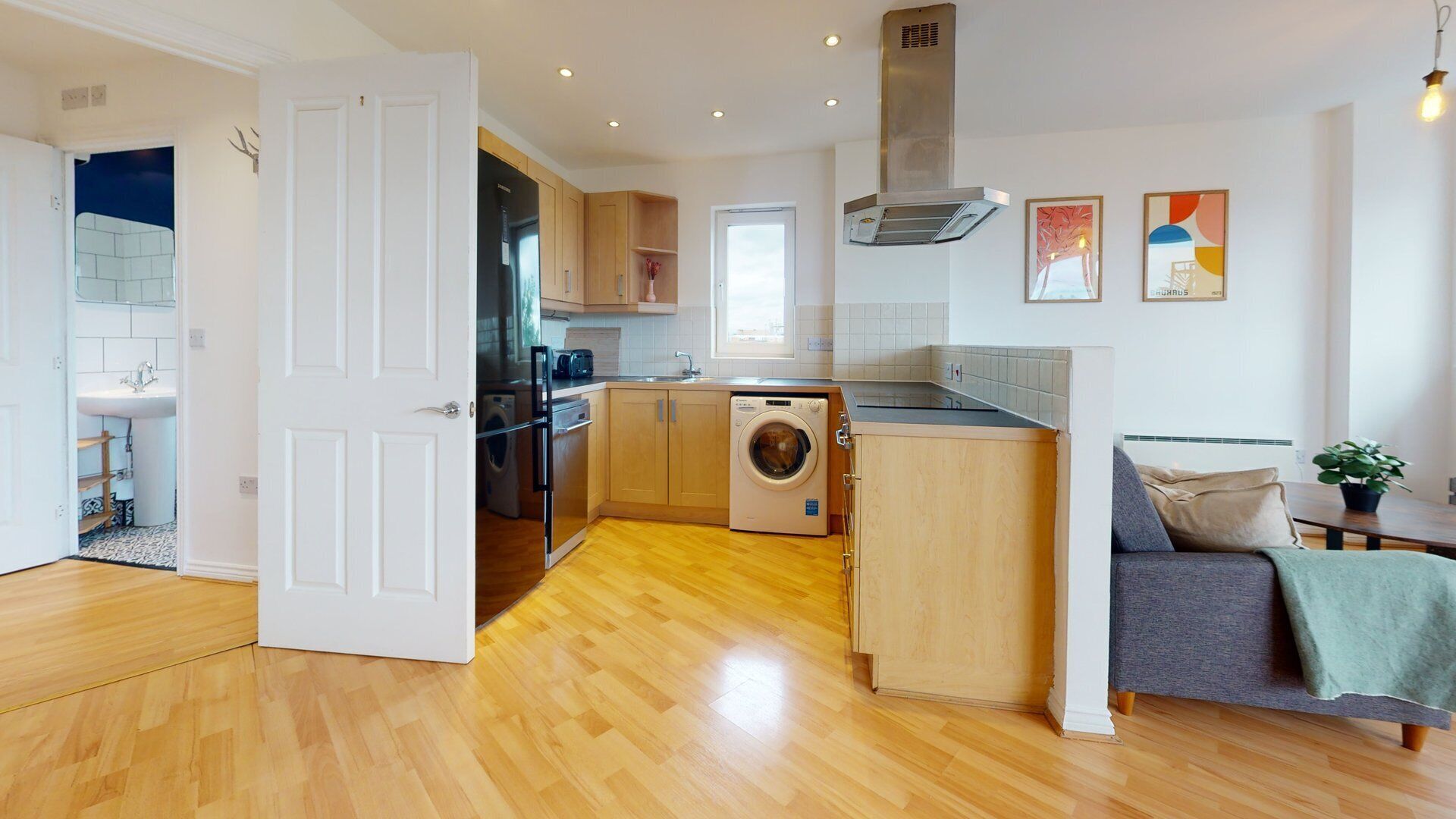 A modern kitchen with wooden cabinets, a built-in washing machine, and a dining area with a sofa and table.
