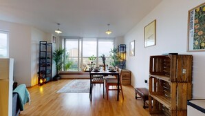 Un espace repas moderne avec une table en bois, des chaises et un banc. On y trouve des plantes en pot, une bibliothèque et une grande fenêtre offrant une vue sur la ville.