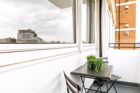 A balcony with a table and chairs, a potted plant, and a view of a building across the street.