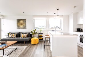 A modern apartment with a grey sofa, yellow ottoman, kitchen island, and a city view.