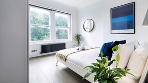 A modern bedroom with a large window, a bed with white bedding, a round mirror, and a framed artwork on the wall. - Modern and Bright Highgate Gem (London)