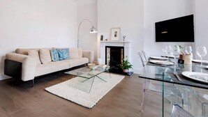 A modern living room with a glass coffee table, a beige sofa, a fireplace, and a dining area with clear chairs and a glass table. - Modern and Bright Highgate Gem (London)
