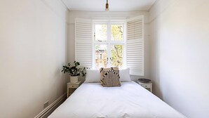 A bedroom with a large bed, white walls, a window with shutters, a bedside table with a plant, and a nightstand. - Stunning Maida Vale Apartment (London)