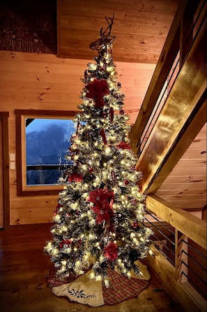A Christmas tree decorated with lights and ornaments, placed in a wooden room with a window overlooking a body of water. - Luxury Cabin, Incredible View, Amazing Sunrise & Sunset, Secluded, Pet Friendly (Blue Ridge)