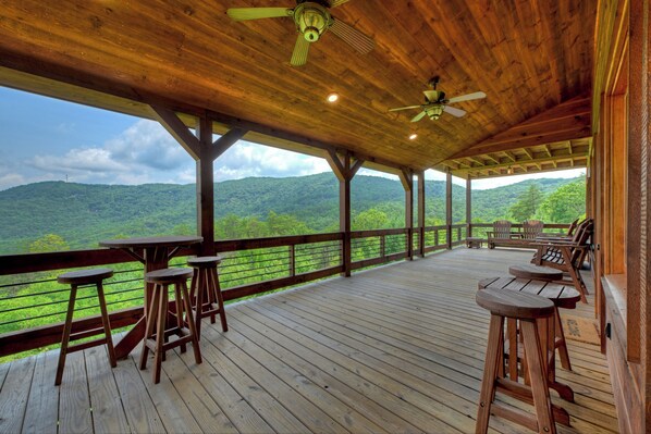 Terraza de madera con vistas a una cadena montañosa boscosa, mobiliario exterior y ventiladores de techo.