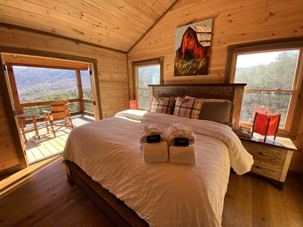 A wooden cabin bedroom with a large bed, a painting above the bed, and a balcony with a view of the mountains. - Luxury Cabin, Incredible View, Amazing Sunrise & Sunset, Secluded, Pet Friendly (Blue Ridge)