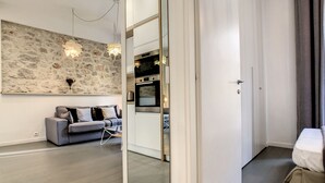 Ein modernes Wohnzimmer mit einer Steinwand, einem grauen Sofa und einer durch eine Glastür sichtbaren Küche.