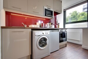 A compact kitchen with a washing machine, a microwave, and a window with a view of trees. - (189OC-9) Cozy Studio Near Beach & Town Centre (Bournemouth)