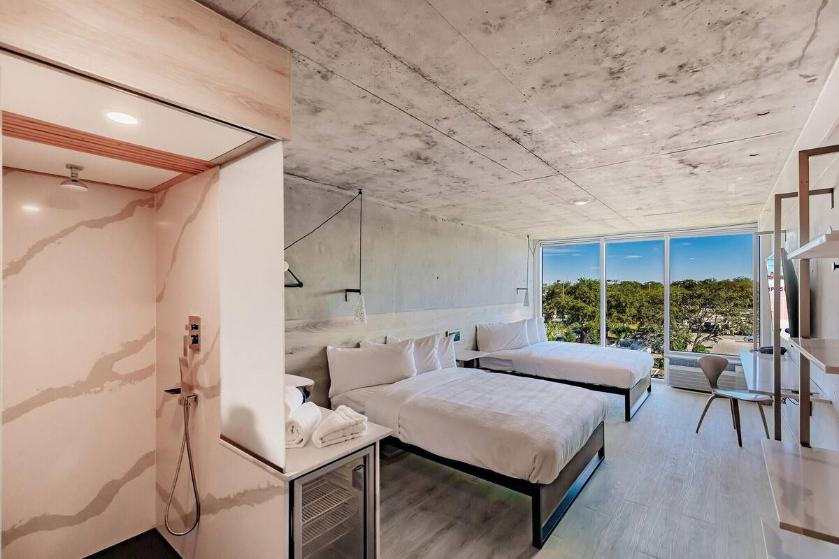 A modern hotel room with a large window, a shower, two beds, and a desk.