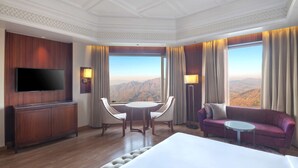 A hotel room with a large bed, a TV, a wooden cabinet, a dining table with chairs, a sofa, and a view of mountains through large windows. - Radisson Hotel Kufri Shimla (Theog)