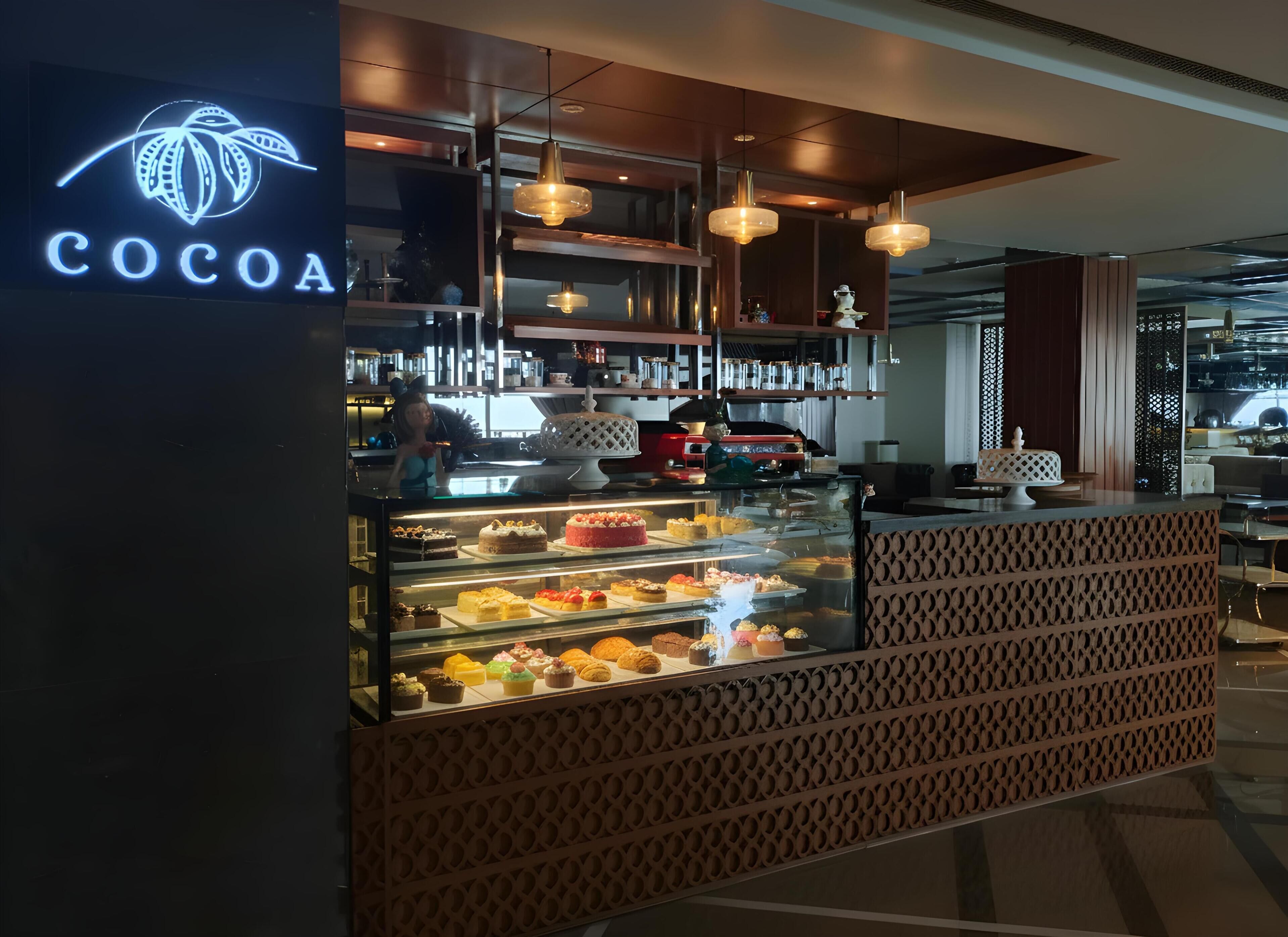 A chocolate-themed dessert display with a variety of cakes and pastries, a modern interior with hanging pendant lights, and a visible kitchen area in the background.