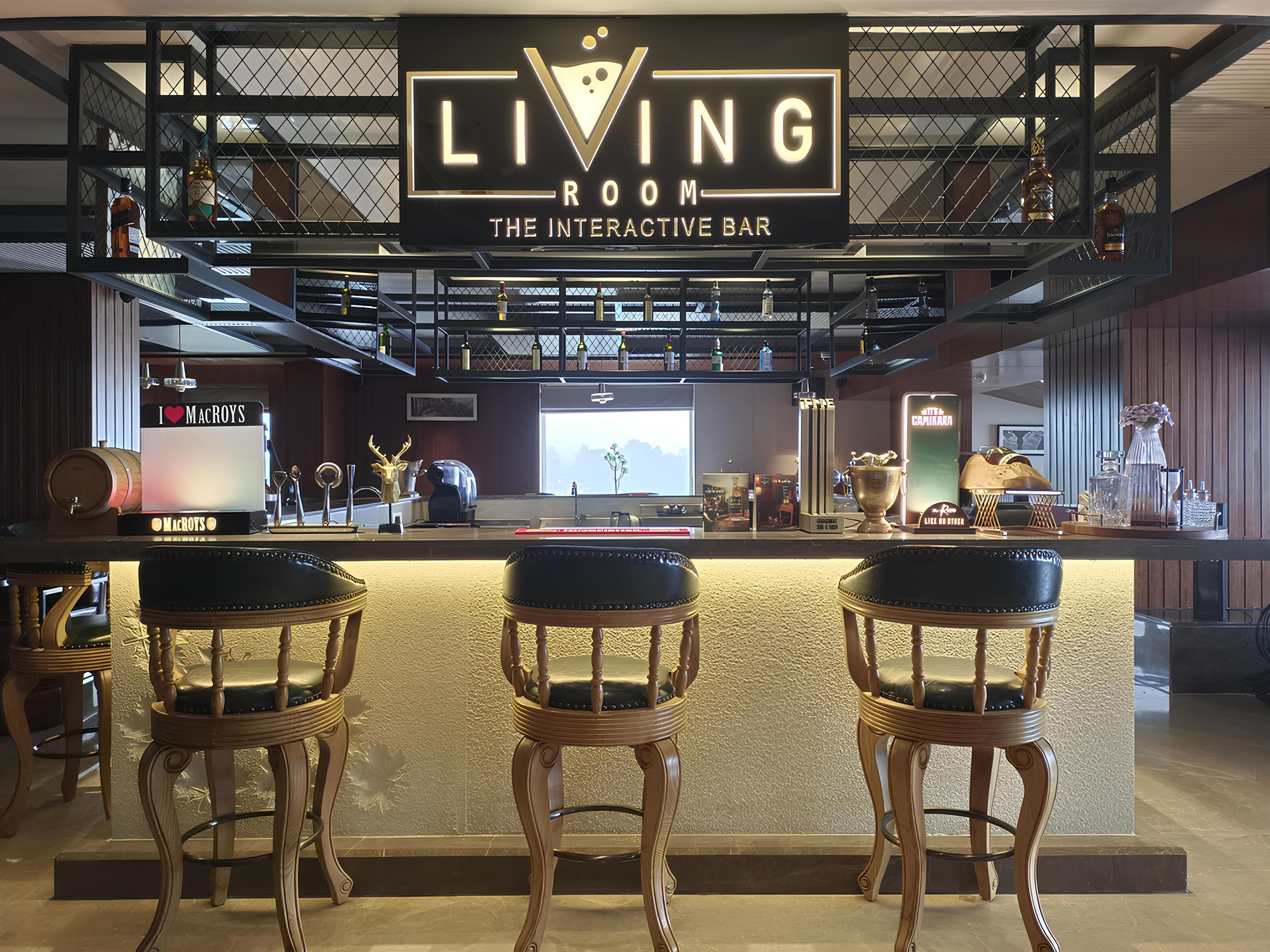 Un espace bar agrémenté d’une enseigne indiquant « LIVING ROOM THE INTERACTIVE BAR », comprenant un comptoir, trois tabourets de bar en bois et diverses bouteilles et verres disposés sur des étagères.