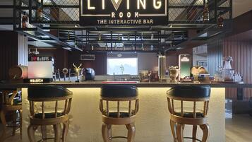 Un espace bar agrémenté d’une enseigne indiquant « LIVING ROOM THE INTERACTIVE BAR », comprenant un comptoir, trois tabourets de bar en bois et diverses bouteilles et verres disposés sur des étagères.