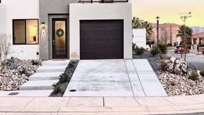 A modern two-story house with a dark garage door, a balcony with a railing, and a small garden area with rocks and plants. - New TownHome w/ Heated Pool & Hot Tub in St.George (St. George)