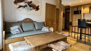 A cozy living area with a sofa, a wooden coffee table, and a fur-covered ottoman. A painting is on the wall, and a kitchen is visible in the background. - Le Jardin Alpin: Cozy apartment in a luxury residence (Les Deux Alpes)