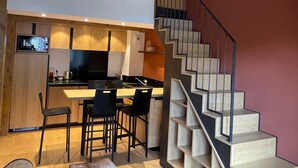 A modern kitchen with a wooden island, black bar stools, and a dining table. A staircase with a unique design leads to the upper floor. - Le Jardin Alpin: Cozy apartment in a luxury residence (Les Deux Alpes)