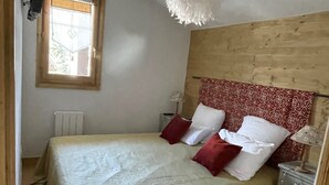 A bedroom with a bed, a wooden headboard, a bedside table, a lamp, and a window with a view of greenery. - Le Jardin Alpin: Cozy apartment in a luxury residence (Les Deux Alpes)