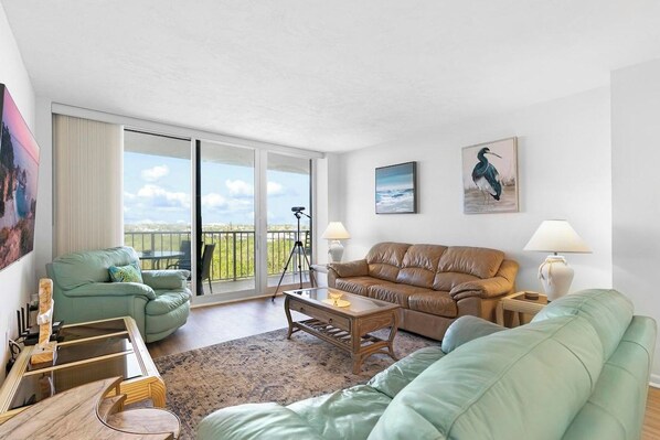 A spacious living room with a large sliding glass door leading to a balcony, a green sofa, a brown sofa, and a wooden coffee table. - South Seas Retreat Beachfront 2BD/2BA- Gulf Views (Marco Island)