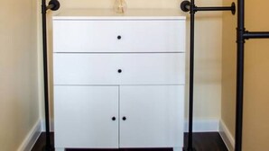 A white dresser with three drawers and a glass vase on top, positioned against a beige wall with a black metal frame structure above it. - Homely Stay Studio3 - with historical mural (München)