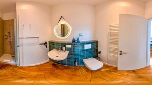 Une salle de bain équipée d’une douche, d’un lavabo et de toilettes, avec un mur recouvert de carreaux verts derrière le lavabo et une porte blanche.
