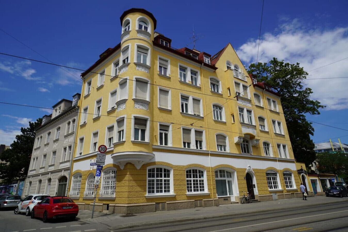 Ein gelbes Gebäude mit zahlreichen Fenstern und Balkonen, ein rotes Auto vor der Fassade und ein Passant, der vorbeigeht.