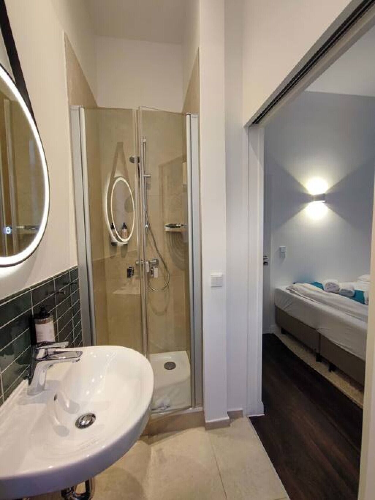 A modern bathroom with a glass-enclosed shower, a white sink, and a bed visible through an open door.