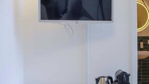 Una habitación moderna con un televisor de pantalla plana montado en la pared, un mini refrigerador blanco con la etiqueta 'CREATE', una mesita de noche blanca con una tetera y un control remoto, y suelo de madera.