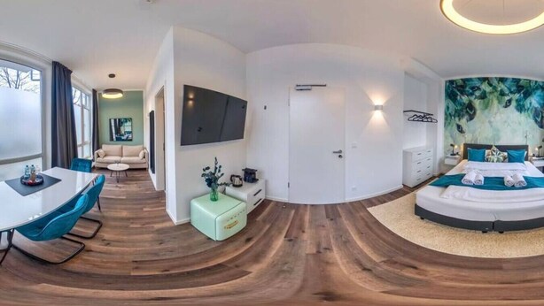 A modern hotel room with a curved wooden floor, a round bed with blue bedding, a flat-screen TV mounted on the wall, and a dining area with a table and chairs. - Homely Stay Studio6 - Modern with feel-good factor (München)