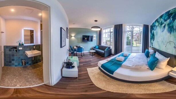Una habitación de hotel moderna con una cama curvada, una zona de estar con sofá y mesa, un baño independiente con lavamanos y un amplio ventanal con vistas.