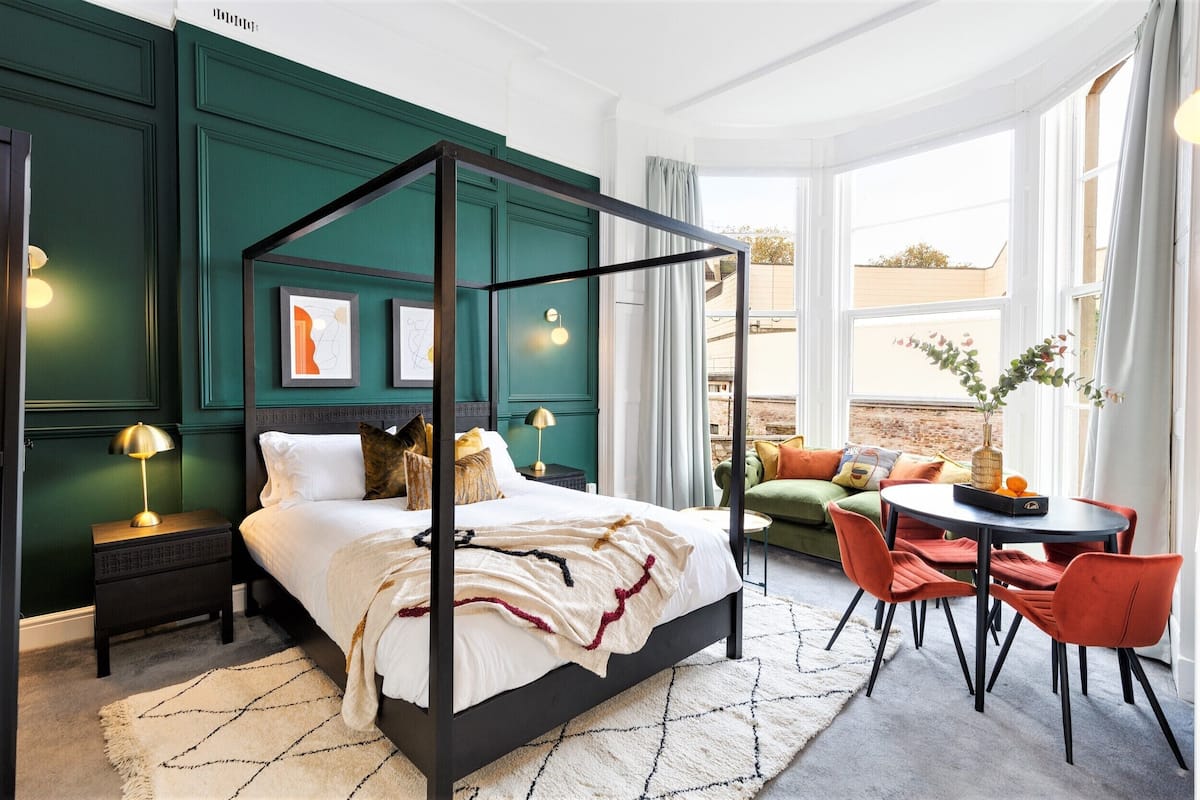 A modern bedroom with a four-poster bed, a green accent wall, and a view of the outdoors through large windows.