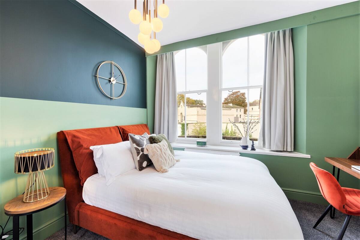 A bedroom with a bed, a round wall clock, a window with curtains, a small table with a lamp, and a chair.