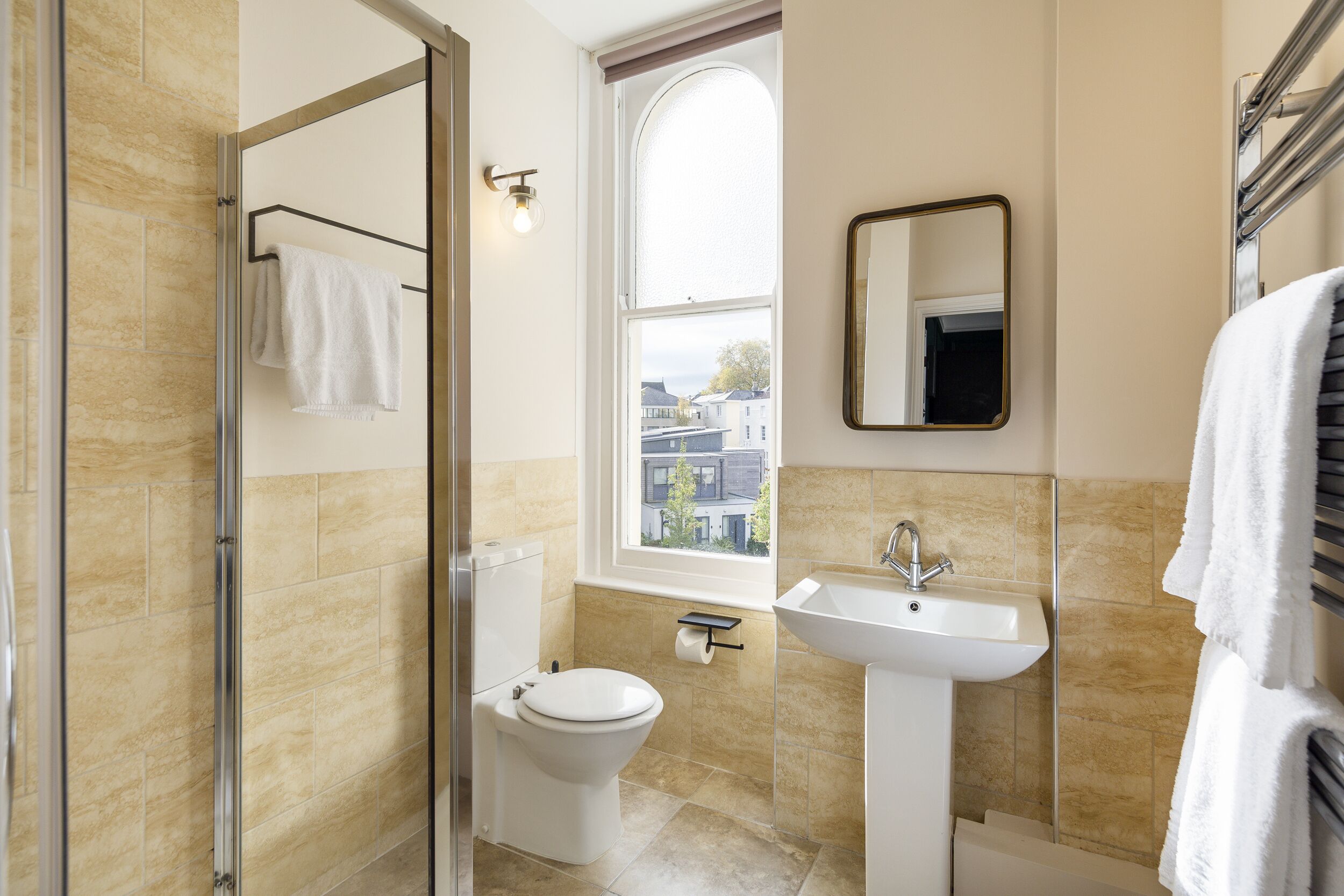 Un baño moderno con cabina de ducha de vidrio, inodoro, lavamanos y ventana con vistas a edificios.