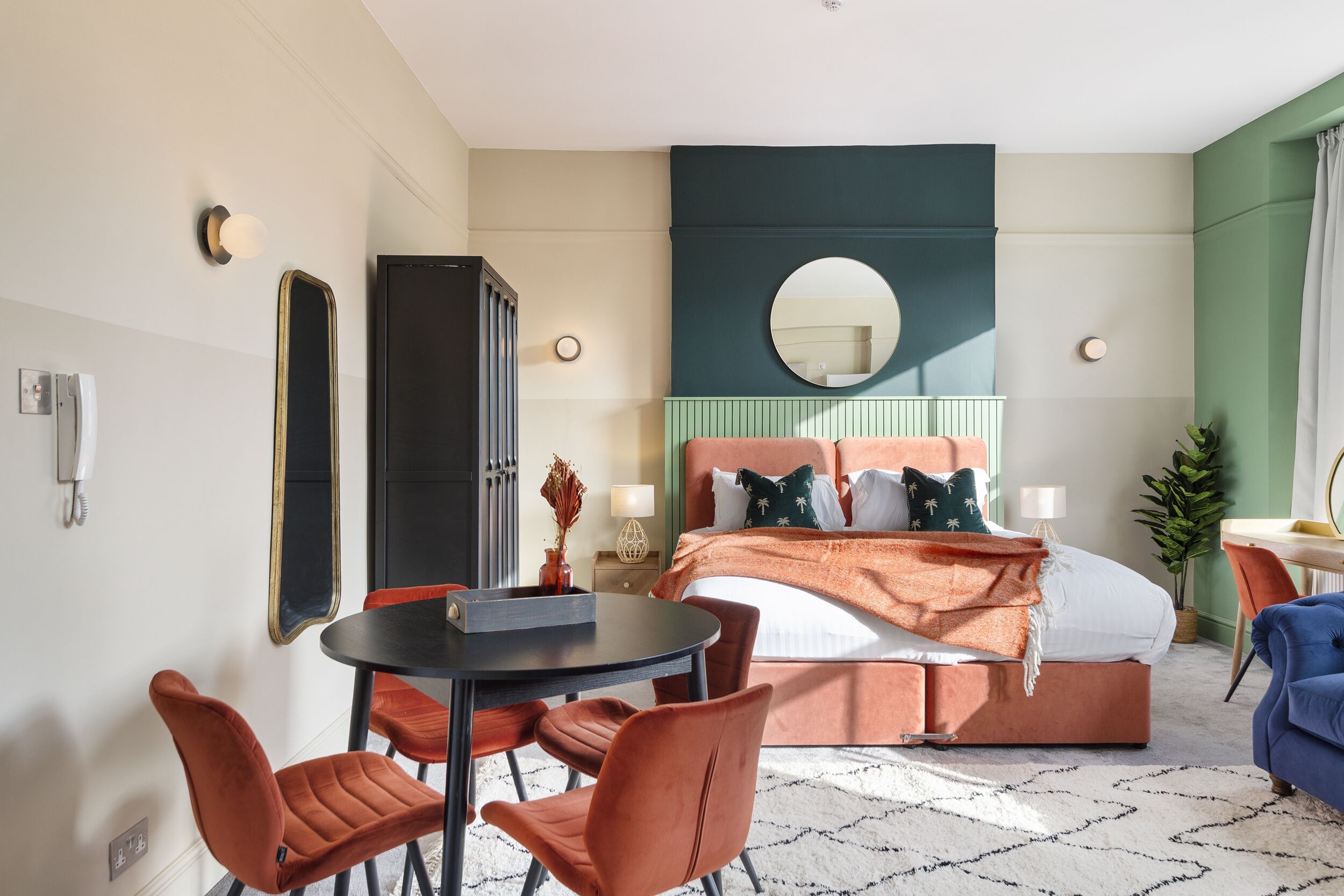 A modern hotel room with a bed, a round mirror, a round table with chairs, and a small desk with a chair.